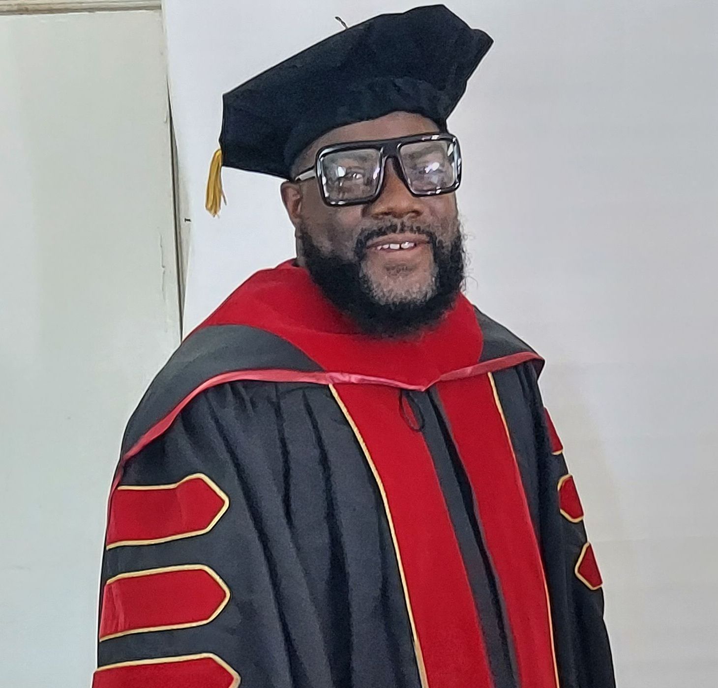 Person wearing academic regalia and cap, smiling. Black robes with red accents, indoor setting.