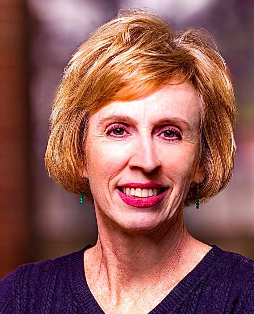 Woman with short blond hair smiles at the viewer, wearing a dark blue shirt; background blurred.