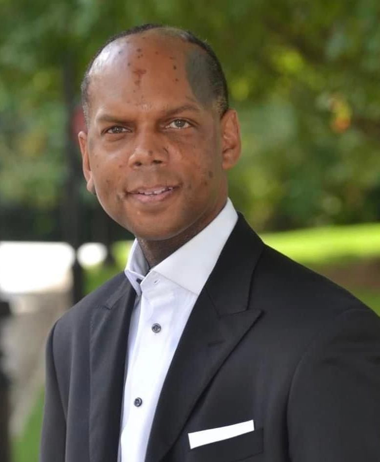 Man in black suit and white shirt smiles outdoors.