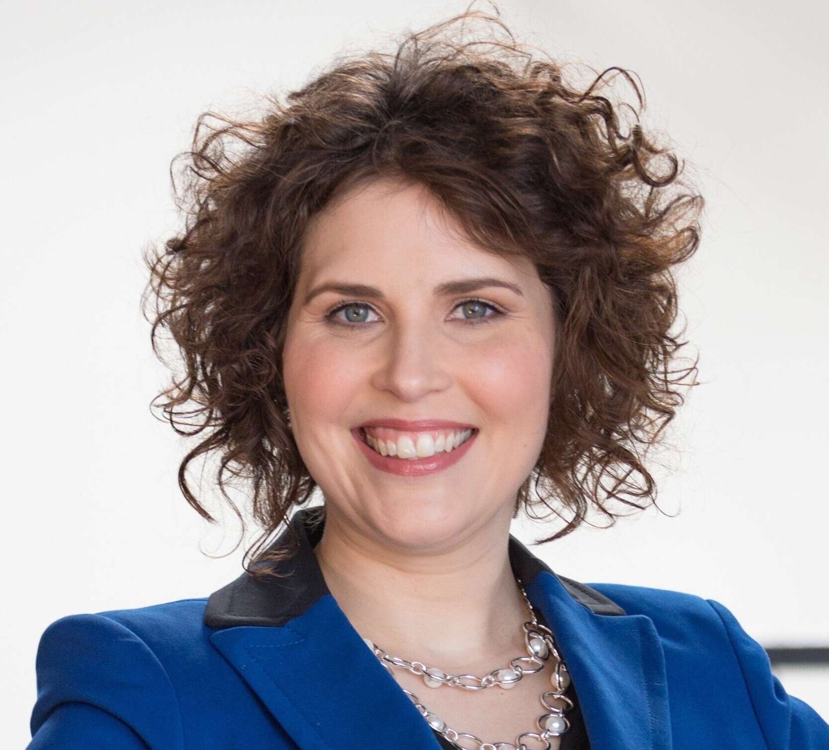 Woman with curly brown hair smiling, wearing a blue blazer and pearl necklace.