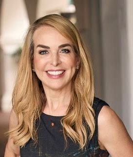 Woman with long blonde hair smiles, wearing a dark top, indoors.