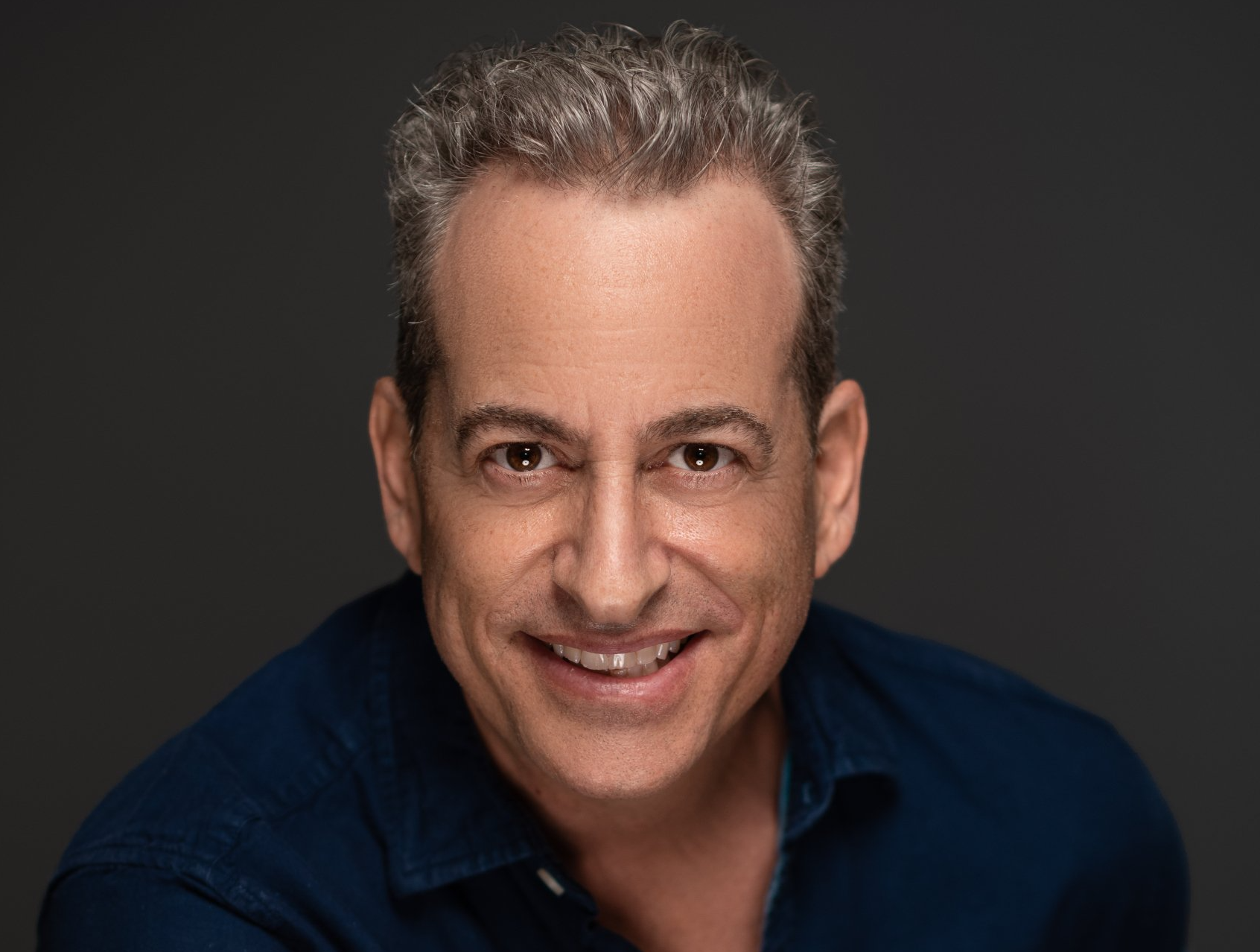 Man smiling, gray hair, navy blue shirt, dark gray background.