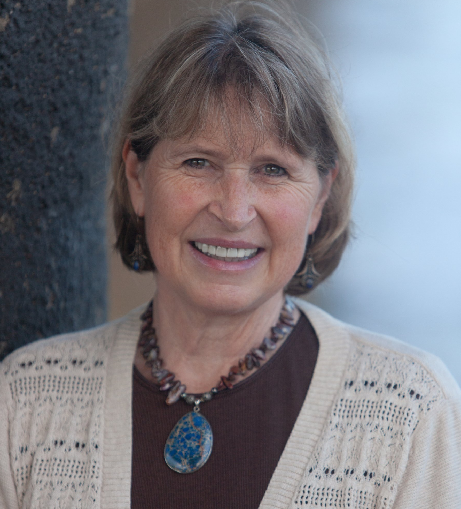 Woman with short brown hair, smiling, wearing a necklace and beige cardigan. Leaning against a dark column.
