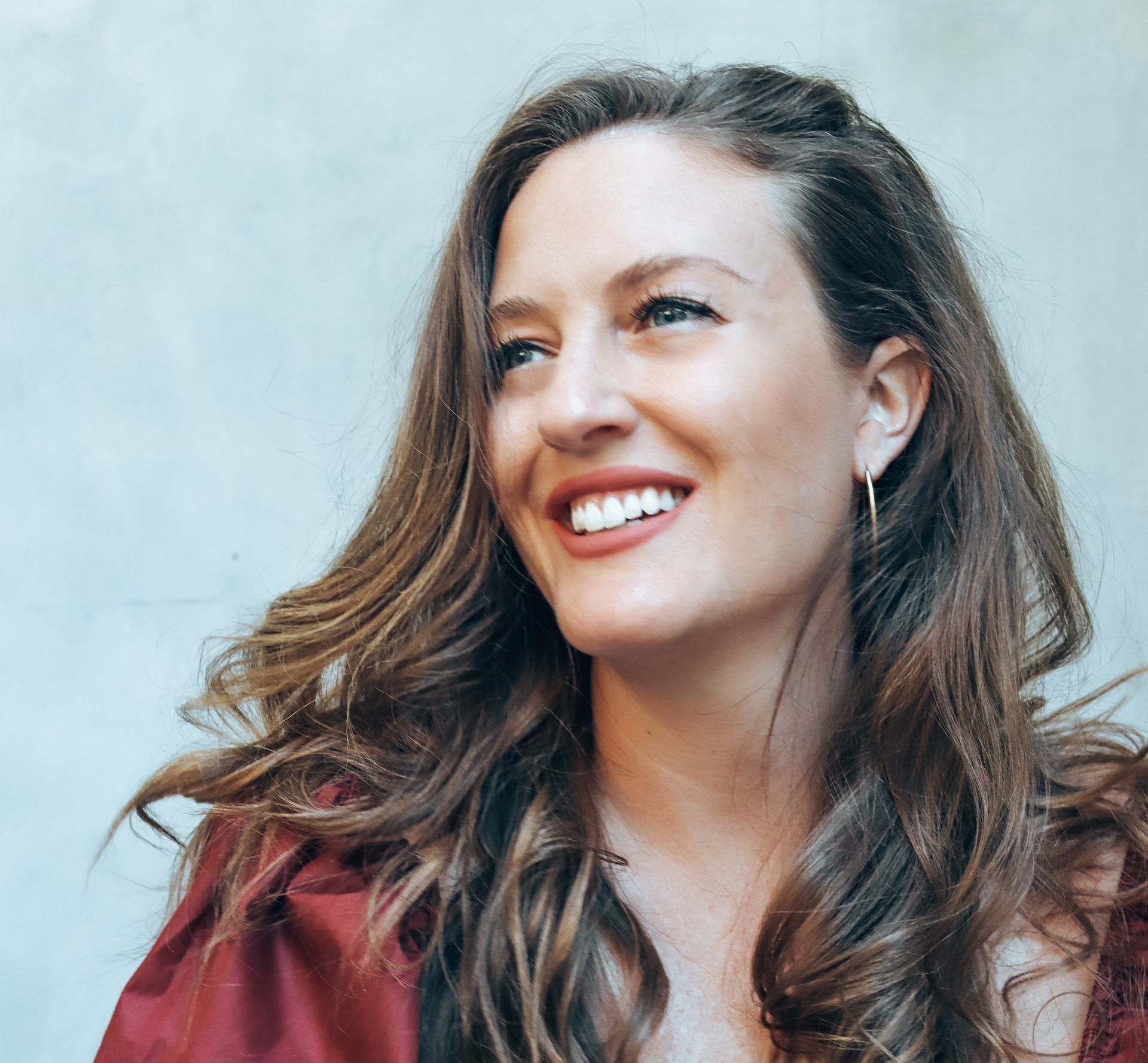 Woman with long, wavy brown hair, smiling with joy, wearing a red top, against a light wall.