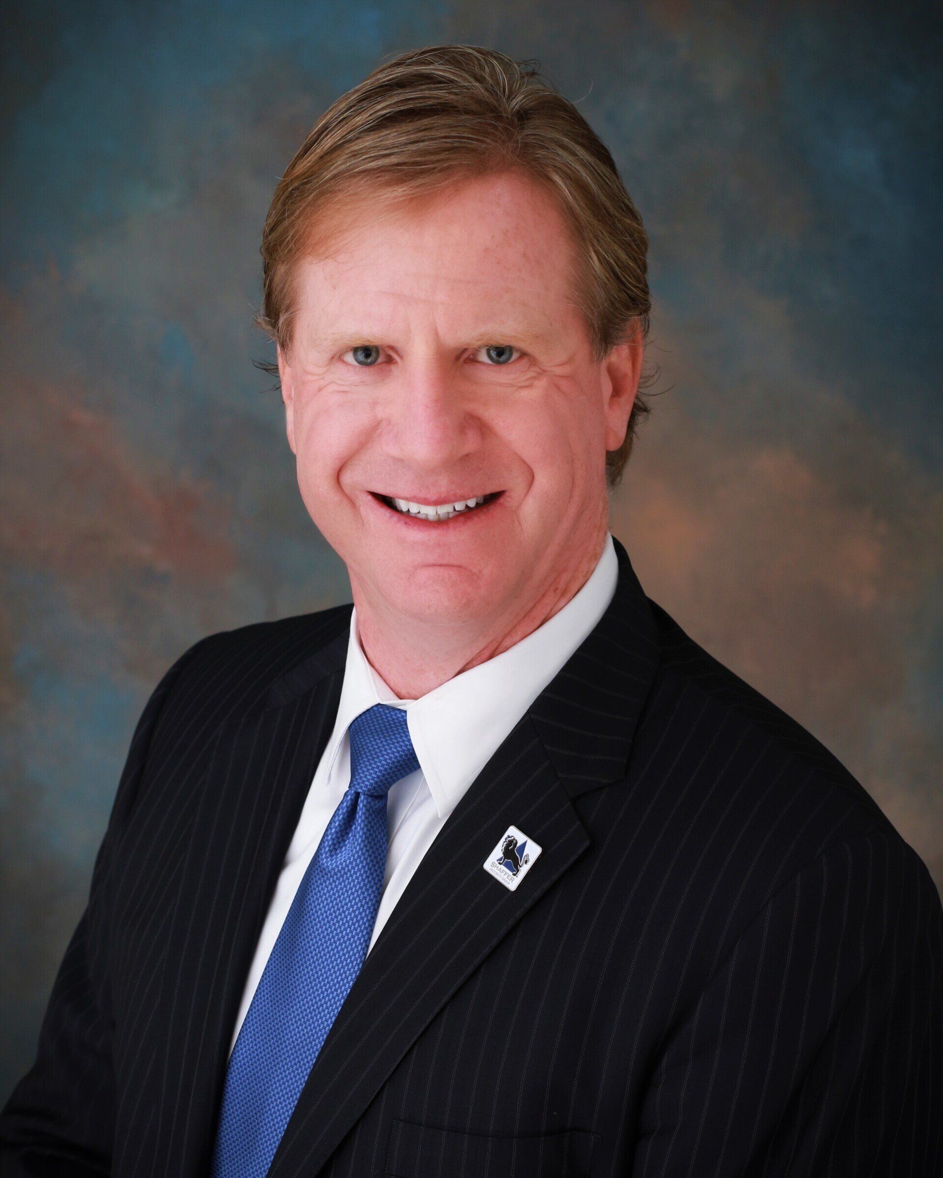 Man in suit smiling, with blue tie and dark blazer, against a blurred background.
