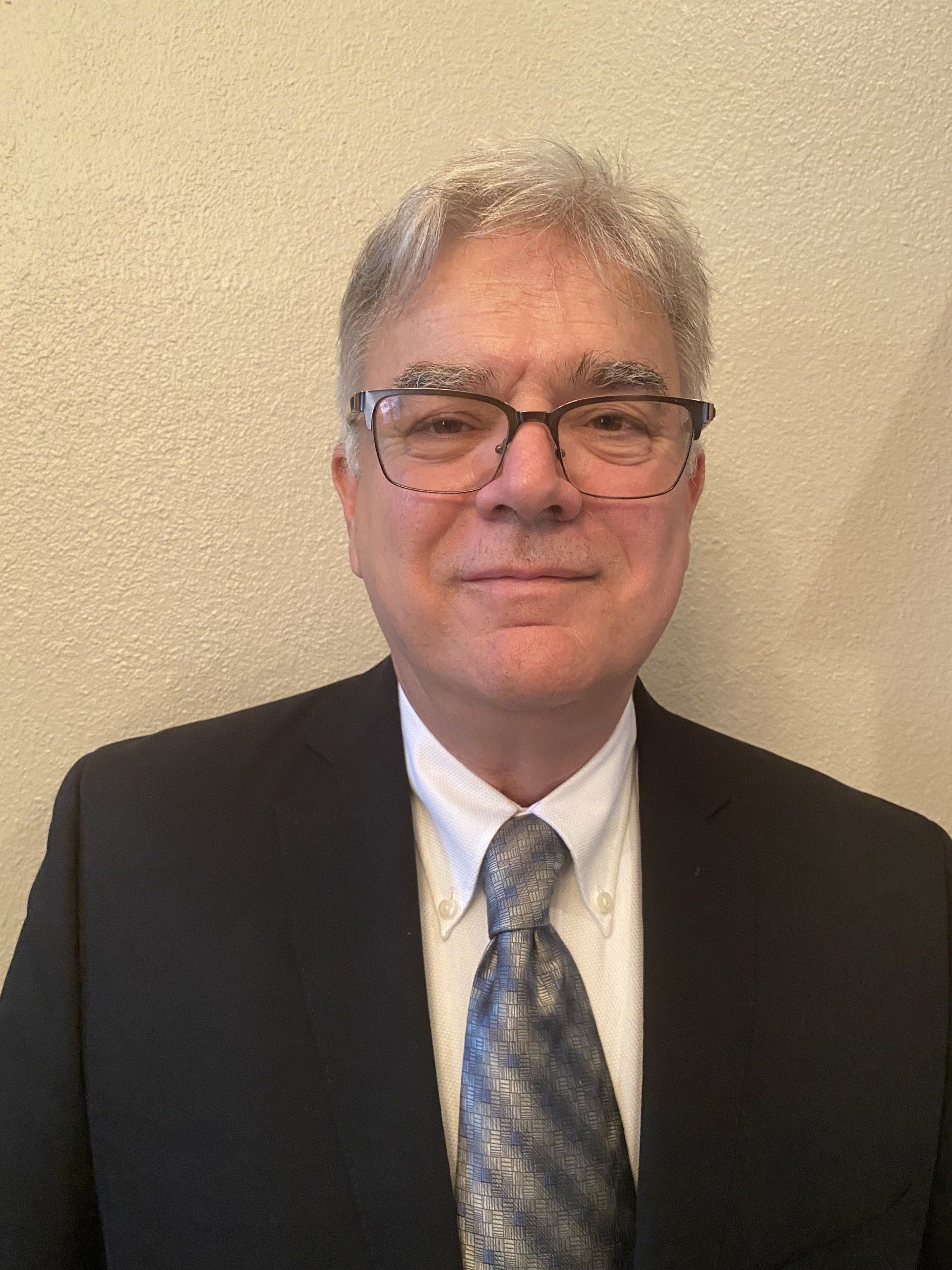 Man in glasses, suit, and tie, standing against a neutral-colored wall.