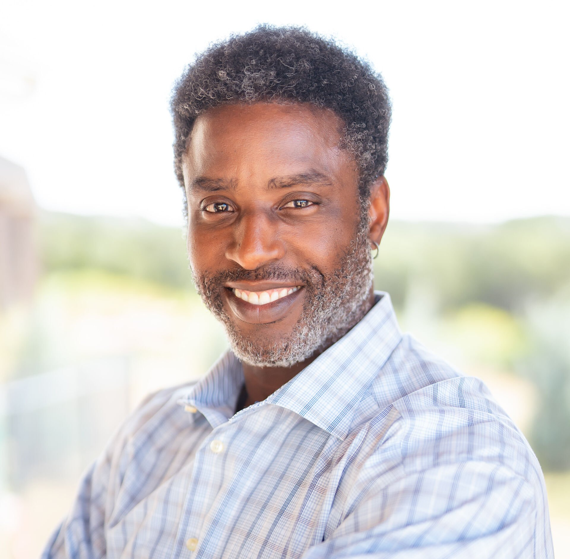 Man with a graying beard smiles, wearing a light blue and white striped shirt, outside with blurred background.