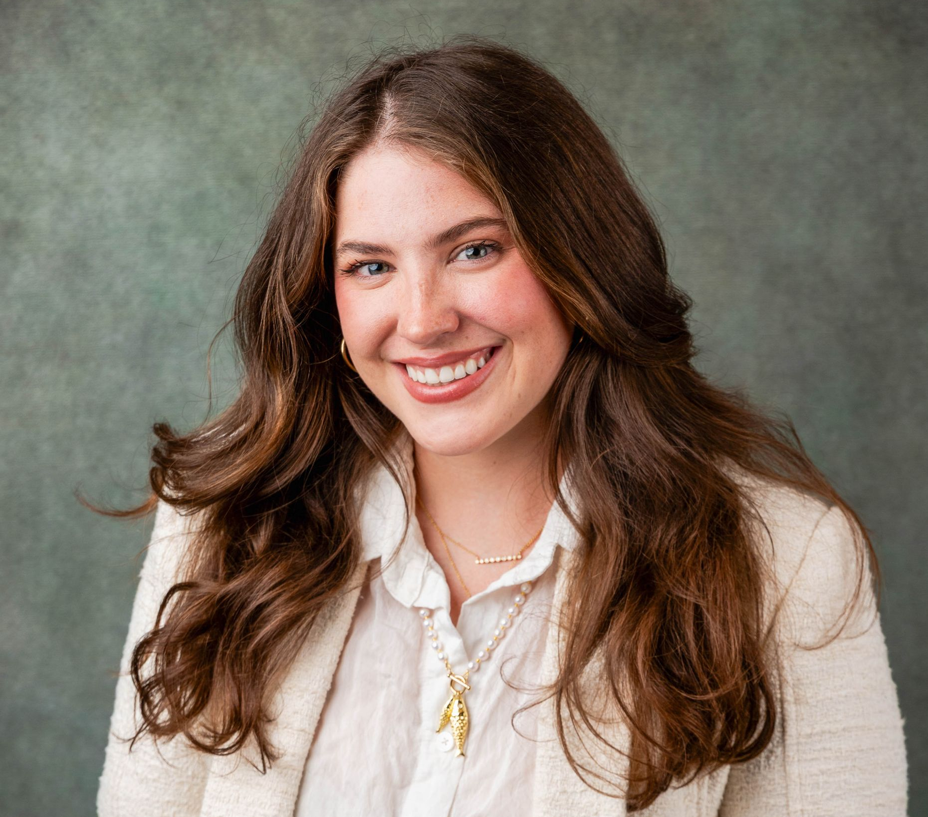 Woman with long brown hair smiles, wearing a white button-down shirt and beige blazer, against a textured green background.