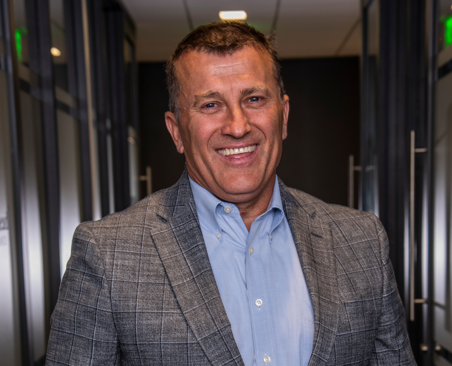 Man in a blue shirt and gray blazer smiles in a hallway.