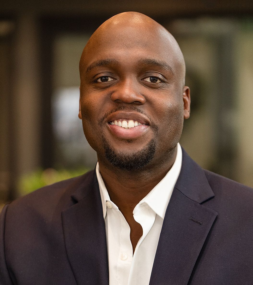 Man with shaved head smiles, wearing a navy suit jacket and white shirt.