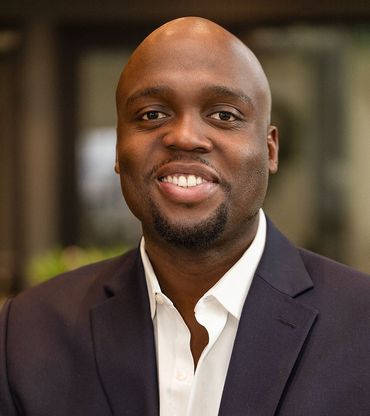 Man with shaved head smiles, wearing a navy suit jacket and white shirt.