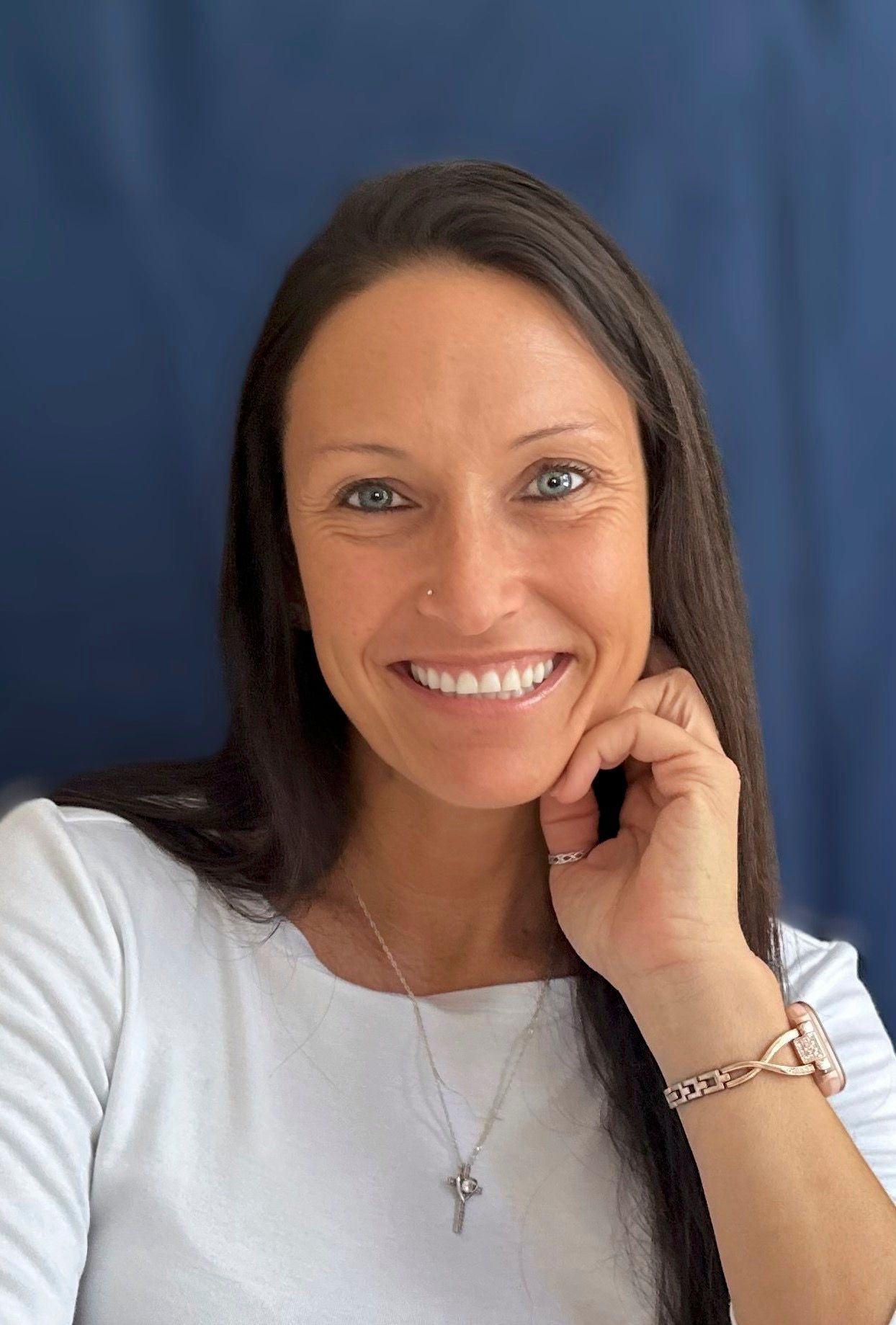 Woman with long dark hair, smiling, resting hand on cheek, wearing a white shirt and necklace.