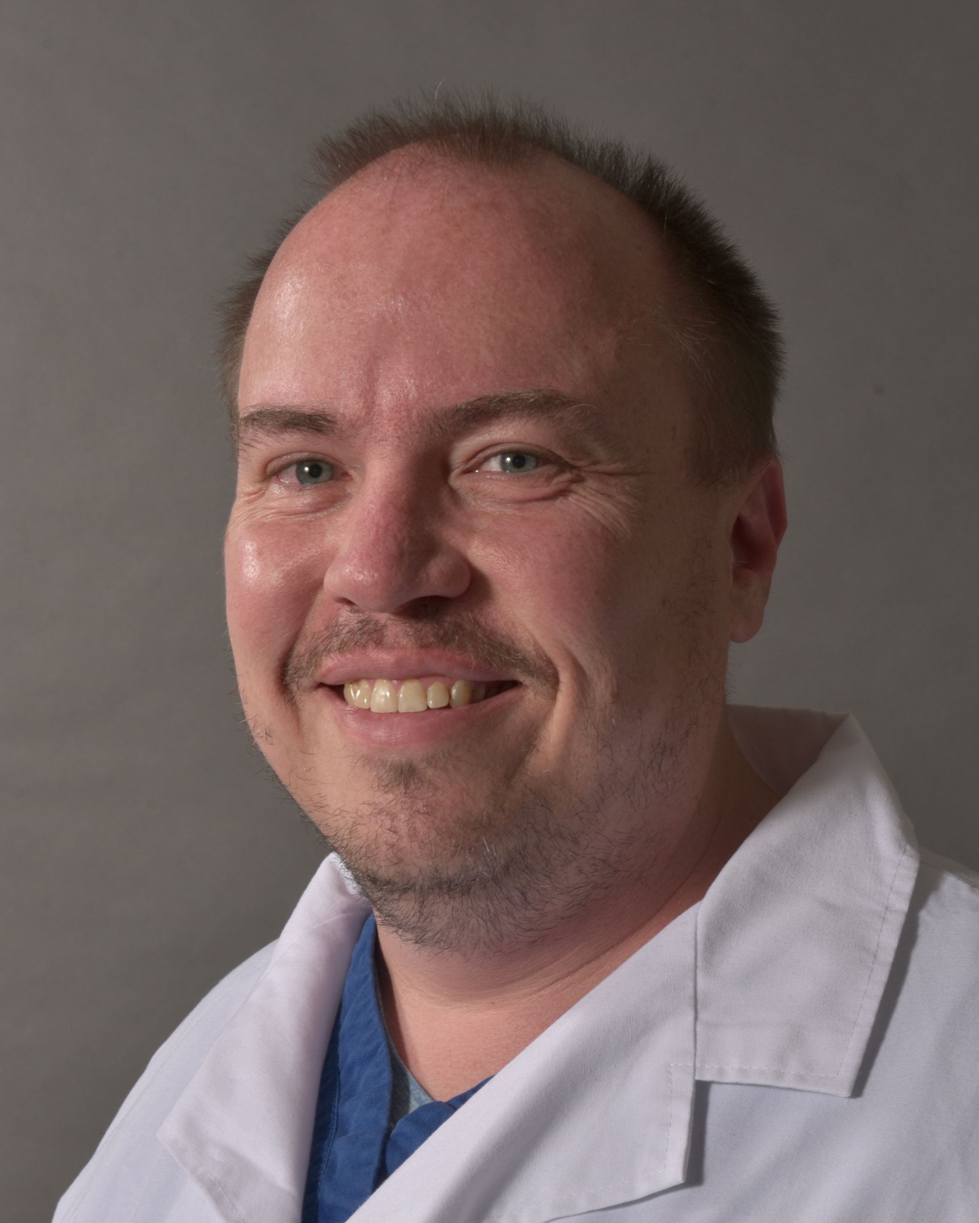 Man in white coat smiles, light skin, short hair, gray background.