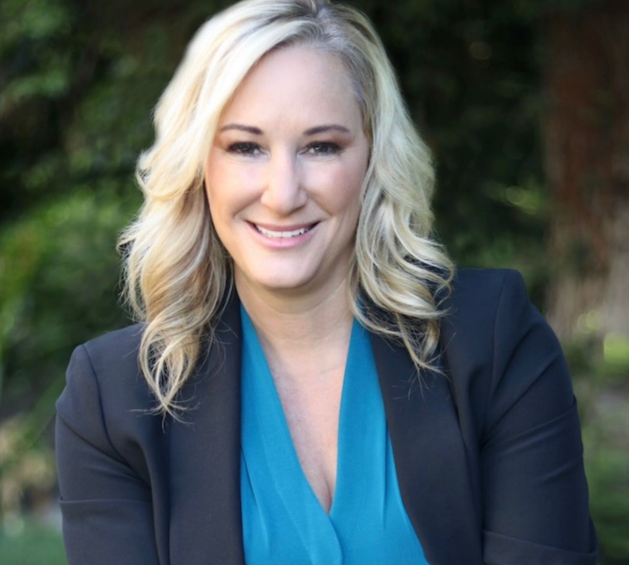 Woman in a teal top and black blazer smiles, outdoors with blurred greenery.