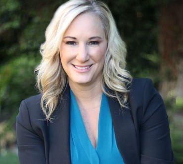 Woman in a teal top and black blazer smiles, outdoors with blurred greenery.