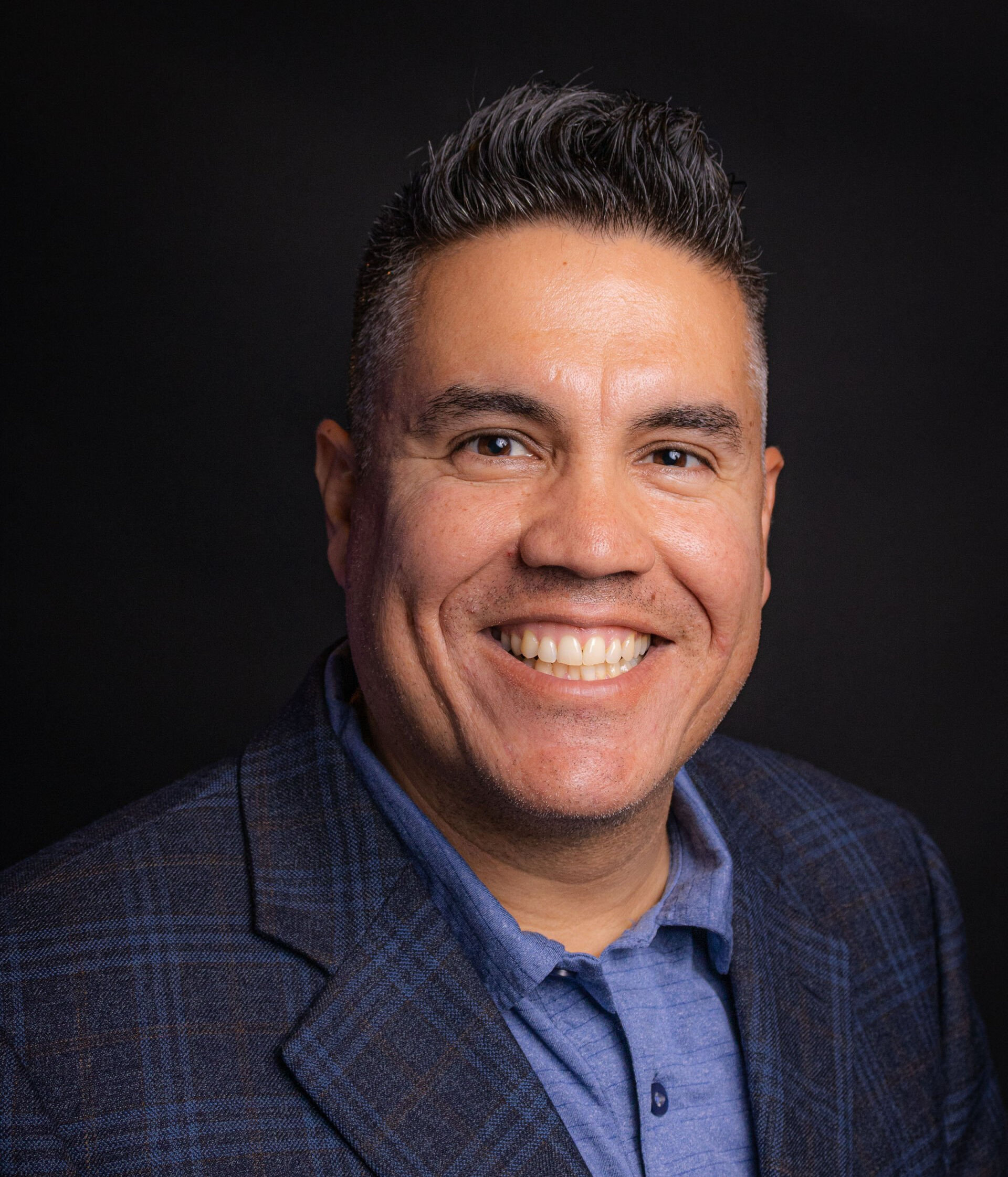 Man with a wide smile, wearing a blue shirt and blazer against a dark background.