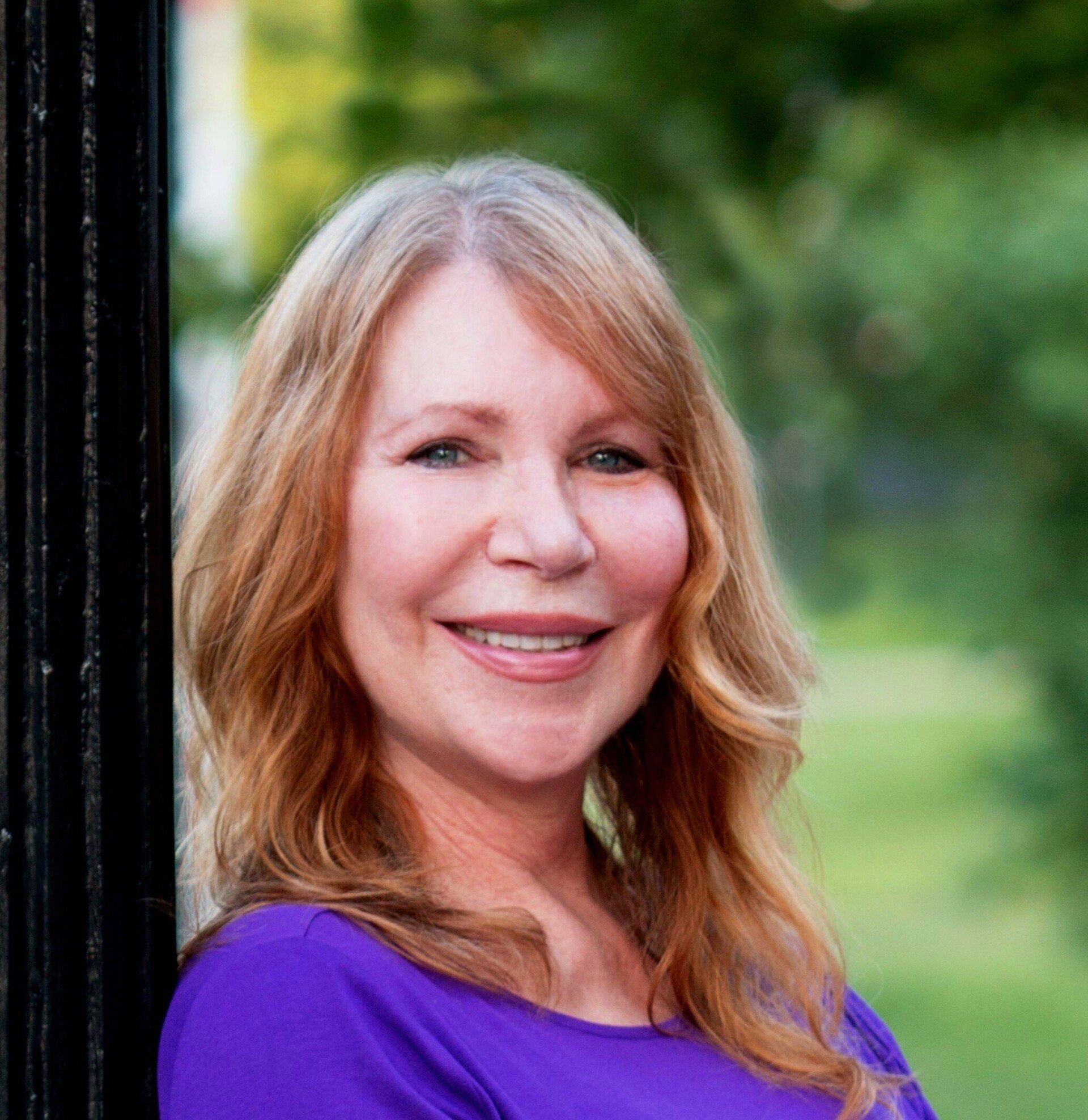 Woman with long, reddish-blonde hair smiles, wearing a purple shirt, leaning on a black fence outdoors.