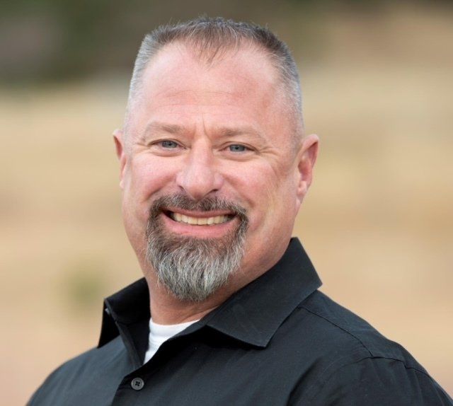 Man with graying goatee smiles, wearing a black shirt with white undershirt, blurred outdoor background.