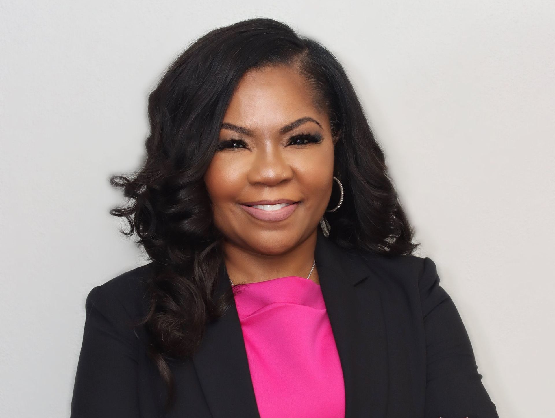 Woman in black blazer, pink top, smiling, against a white backdrop.