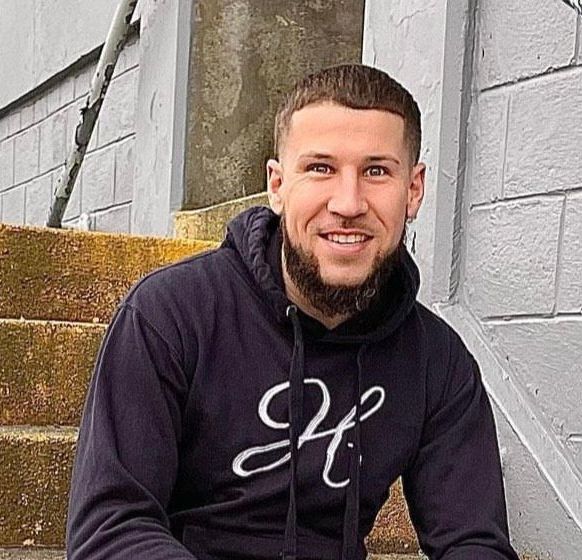 Man in a black hoodie smiles on outdoor stairs.