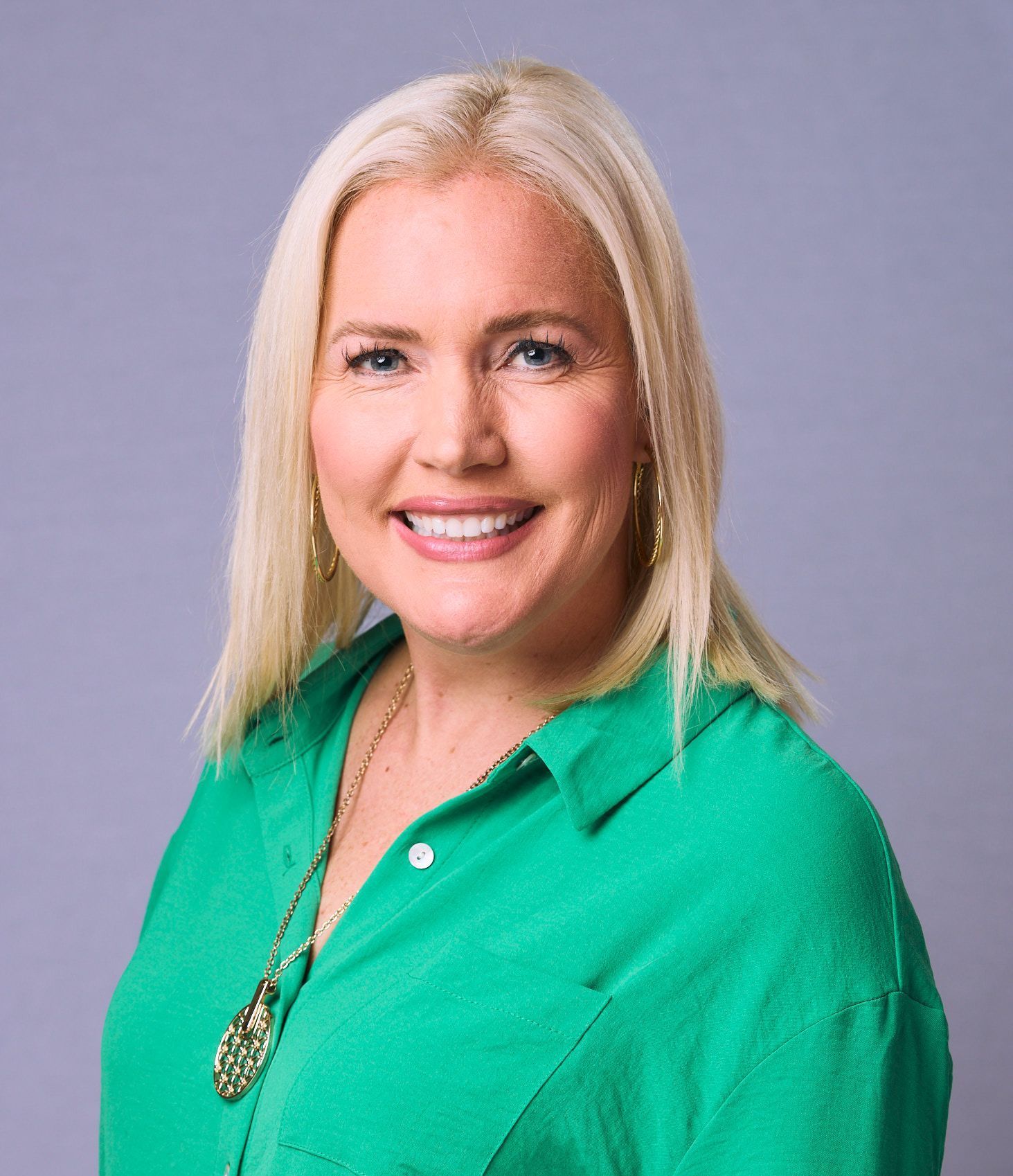 Woman with blonde hair, wearing a green shirt, smiles, facing the camera against a gray backdrop.