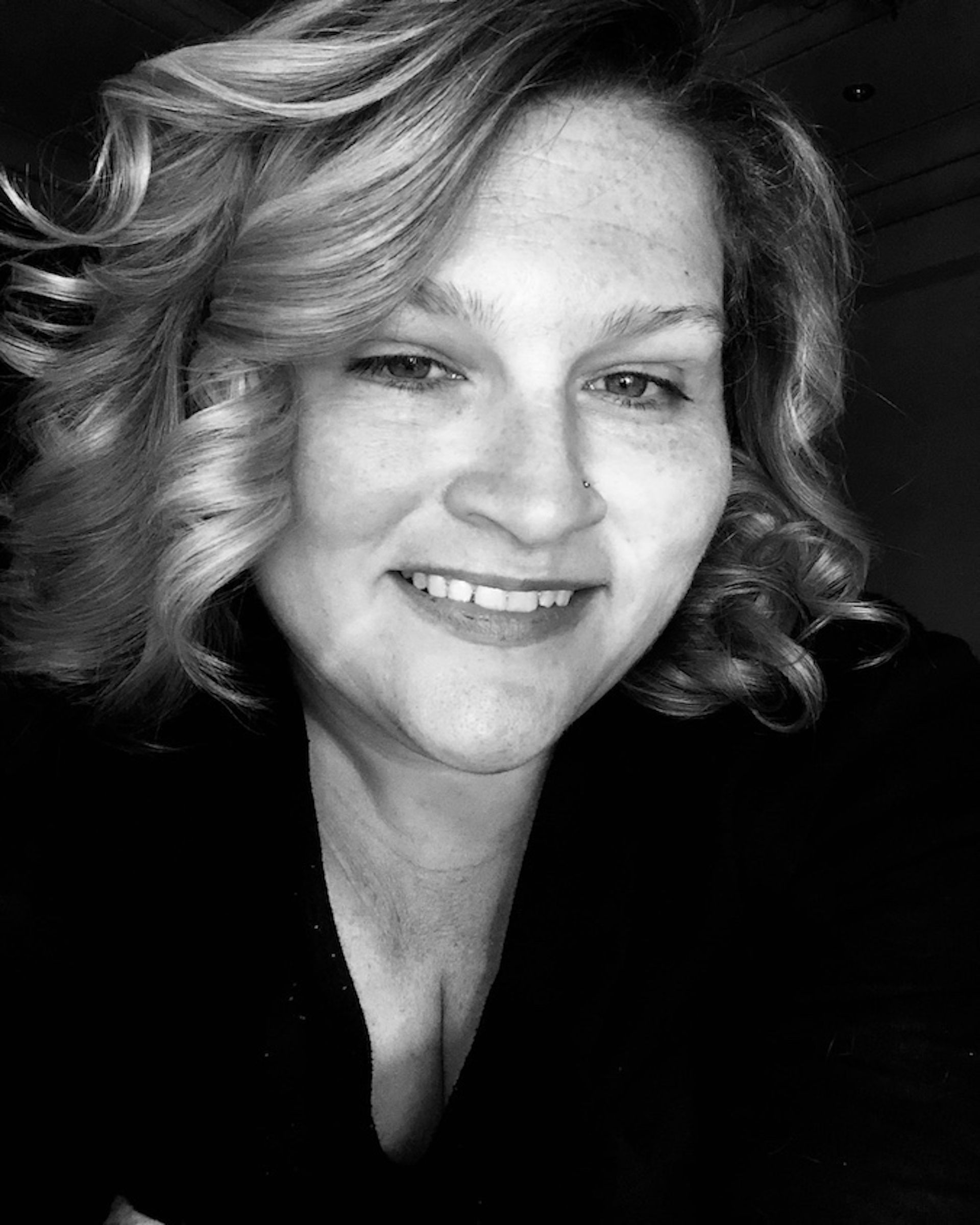 Woman with curly blonde hair smiles, wearing a dark top, in a black and white photo.