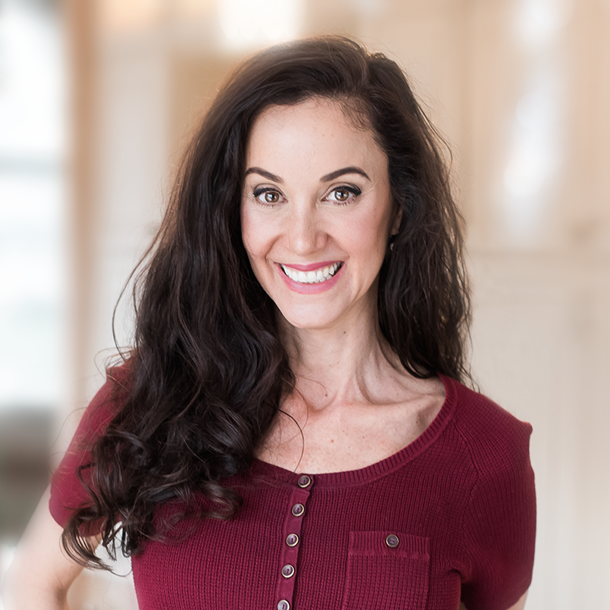 Woman with dark hair smiles, wearing a burgundy shirt, in a bright, blurred background.