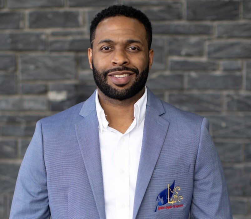 Man in blue blazer smiles, standing in front of a brick wall.