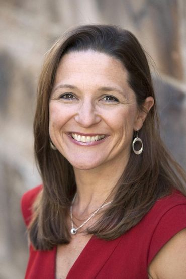 Woman with shoulder-length brown hair smiles, wearing a red top and silver jewelry, in front of a stone wall.