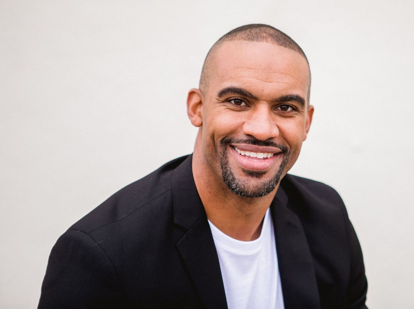 Man in black jacket and white shirt smiling. Background is off-white.