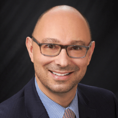 Smiling man with glasses, wearing a blue suit and striped shirt, against a dark backdrop.