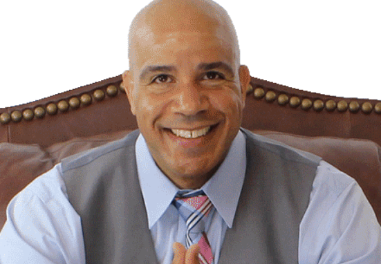 Bald man in gray vest and blue shirt smiles, seated on a leather chair.