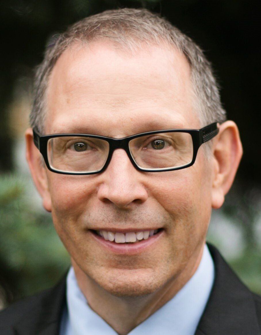 Man in black glasses smiles, wearing a suit jacket and light blue shirt.