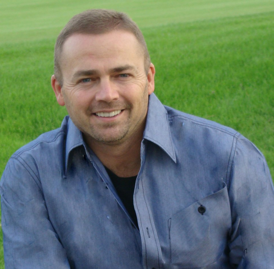 Man with short hair smiles at the camera, wearing a blue shirt, in front of green grass.