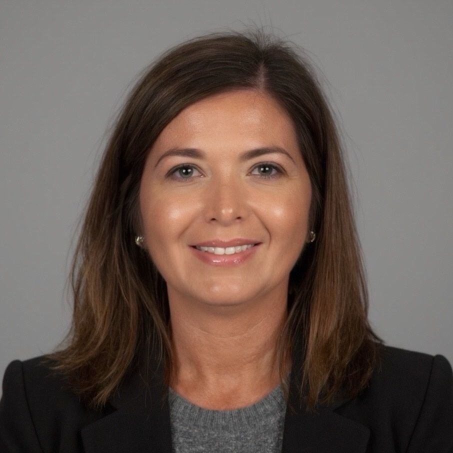 Woman in a black blazer and gray sweater smiles at the camera, against a gray backdrop.