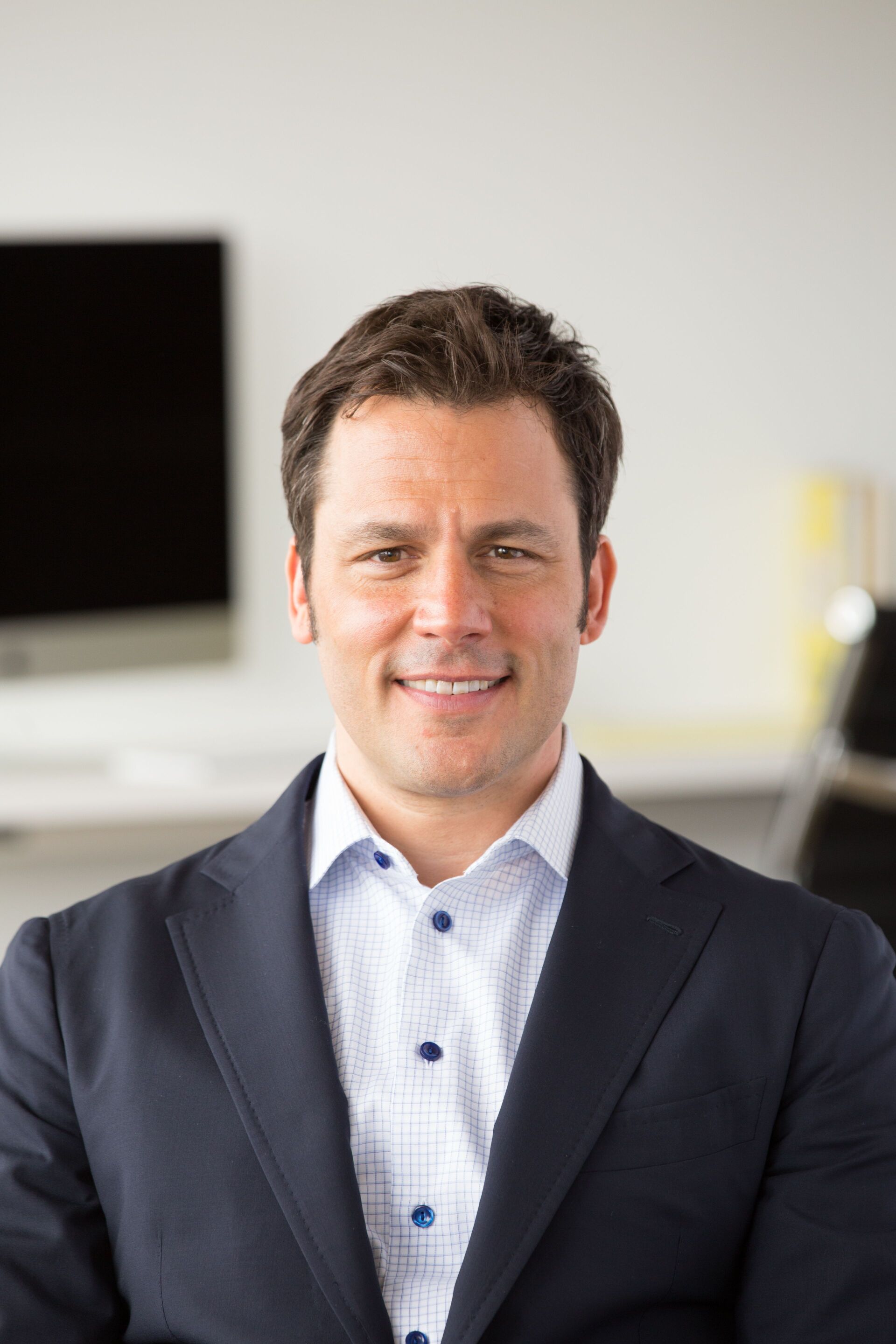 Man in a dark blazer and patterned shirt smiling in an office setting.