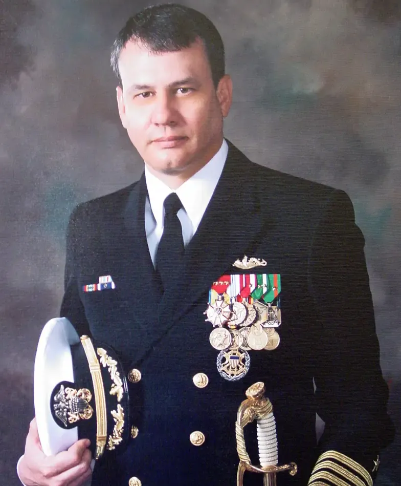 Naval officer in uniform, holding a cap and sword, wearing medals. Dark suit, neutral expression.