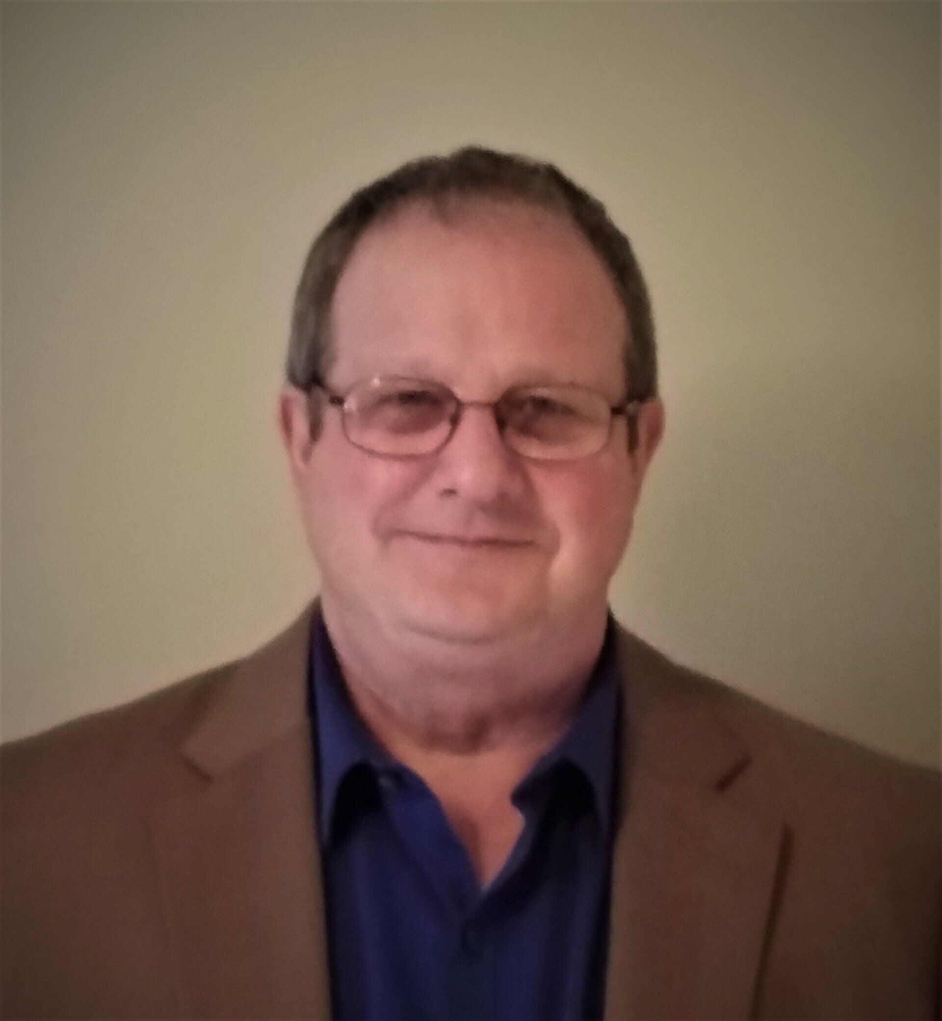 Man wearing glasses, brown blazer, and blue shirt, smiling.