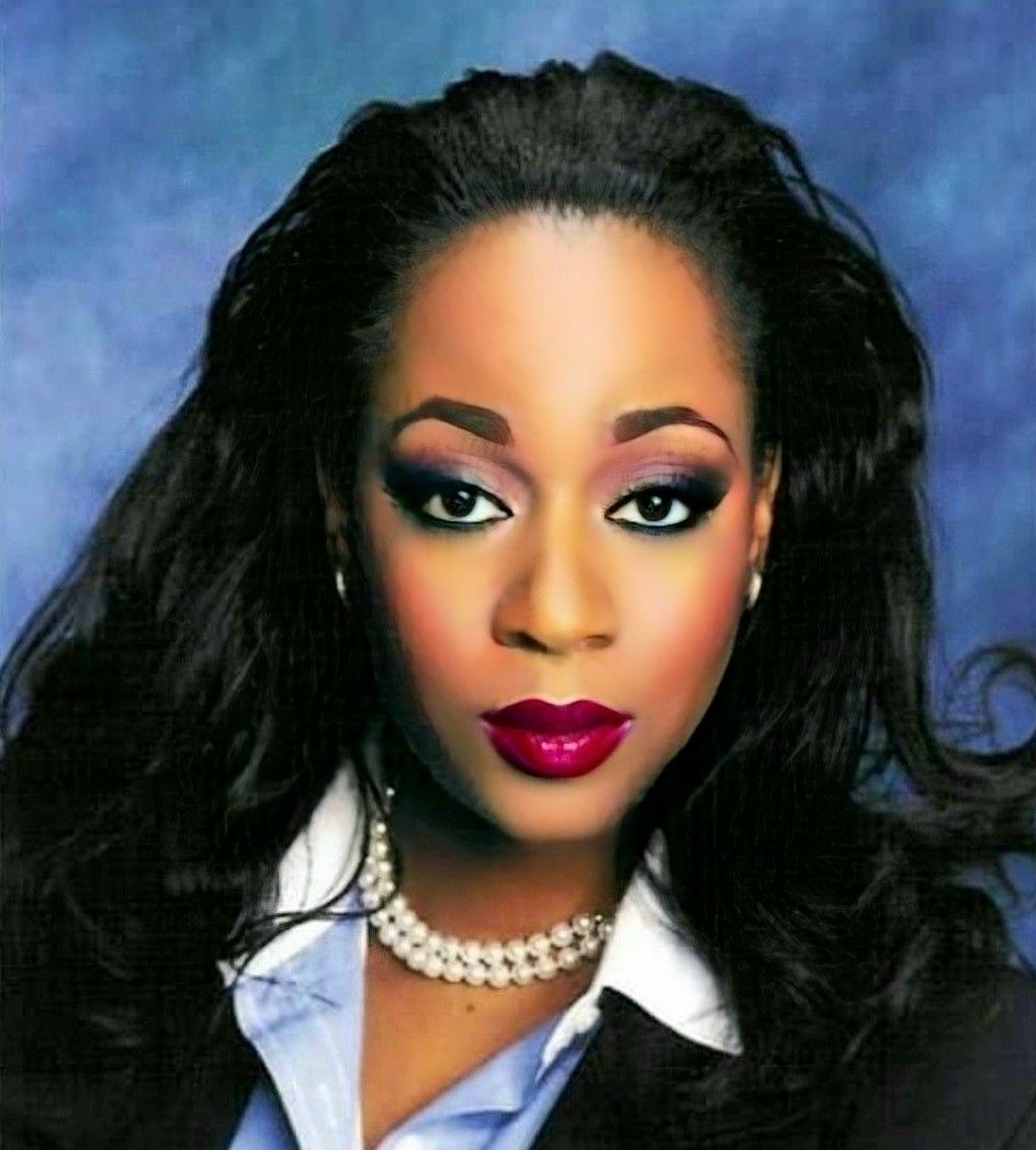 Woman in business attire with dark makeup and pearl necklace.