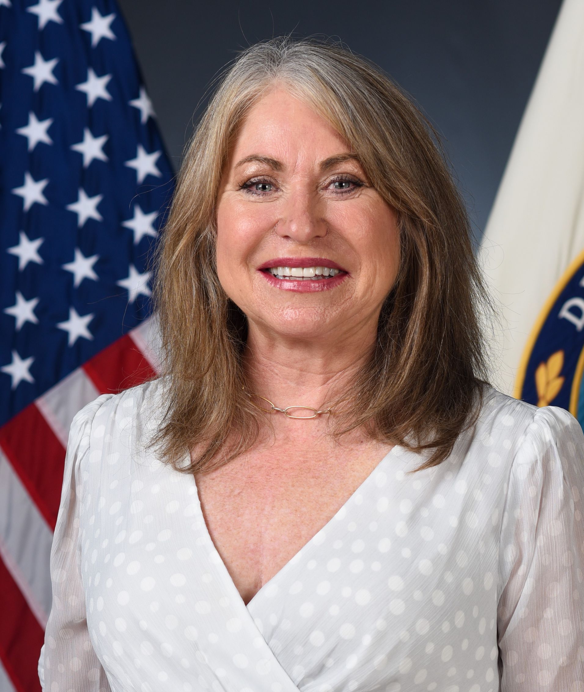 Woman in white top smiles, standing before American flag and Department of Education seal.