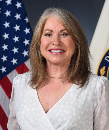 Woman in white top smiles, standing before American flag and Department of Education seal.