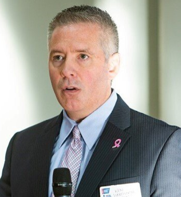 Man in suit speaks into a microphone; wears a pink ribbon pin.