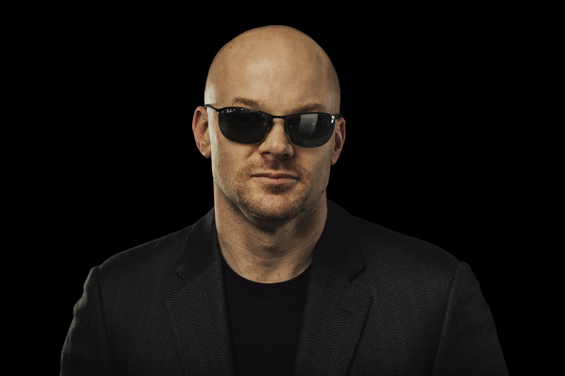 Bald man wearing sunglasses and a blazer, against a black background.
