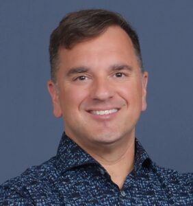 Man smiling, wearing a patterned blue shirt, against a solid blue background.