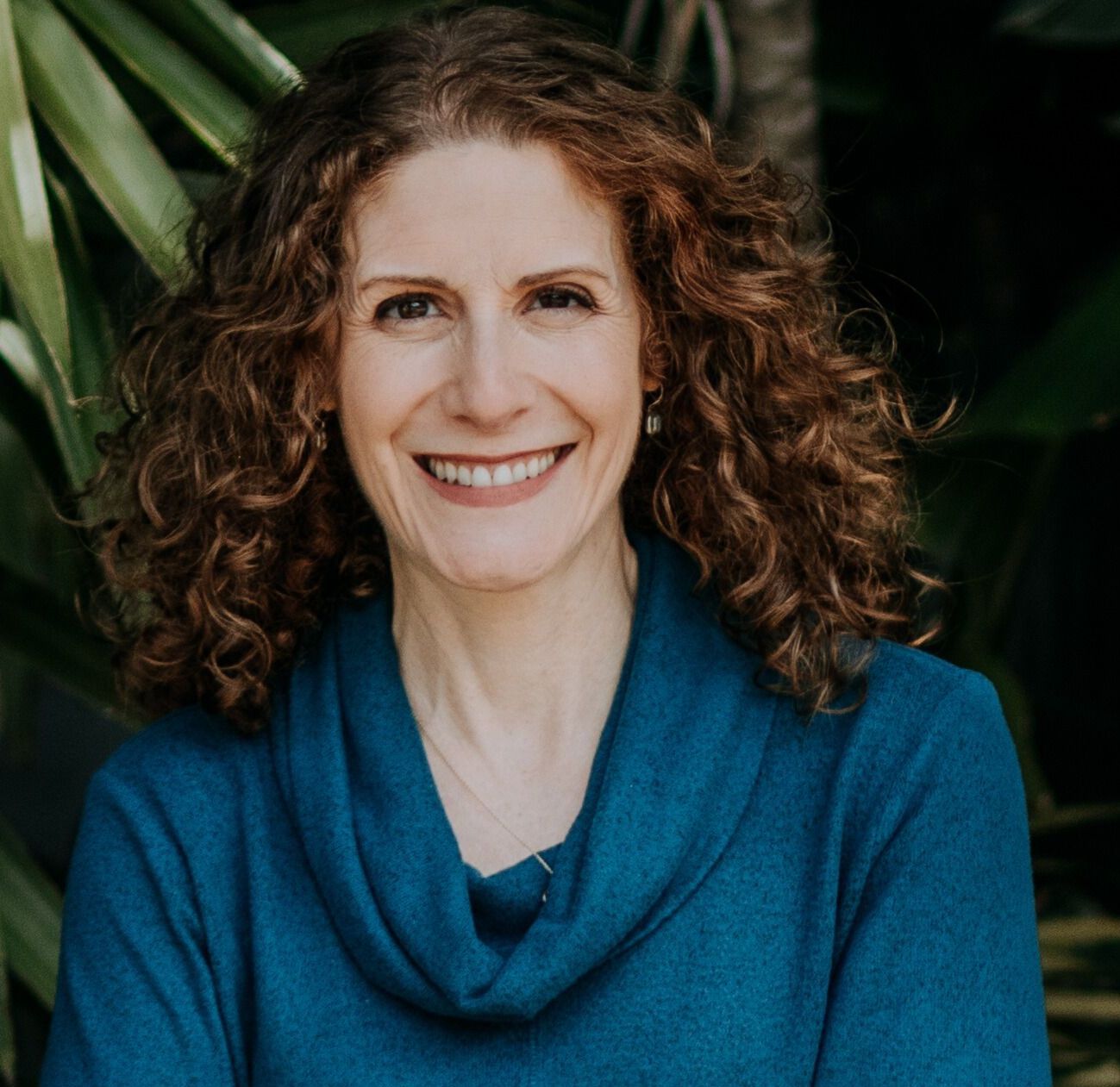 Woman with curly auburn hair smiles, wearing a teal sweater, in front of greenery.