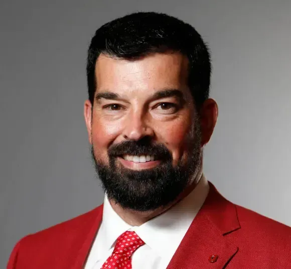 Man in red suit and tie smiles at the camera, against a gray background.