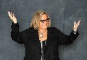 Woman in black blazer, glasses, arms outstretched, smiling, against a gray background.