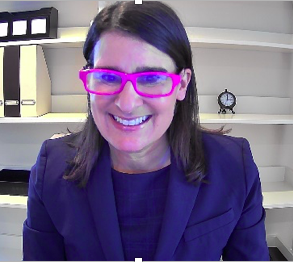 Woman smiling, wearing bright pink glasses and a blue blazer, in a home office with shelves.