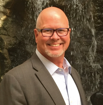 Man in gray blazer smiles in front of a waterfall.