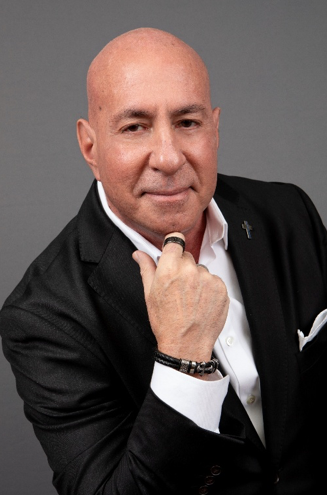 Bald man in a black suit and white shirt, holding a finger up, looking at the camera.
